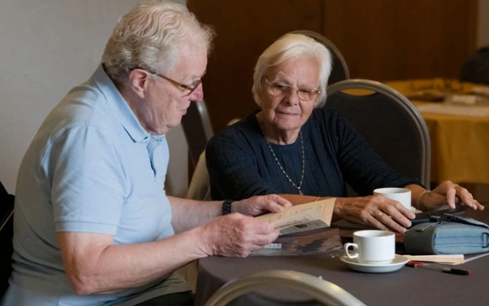 An older couple sitting around a table looking at a book. 